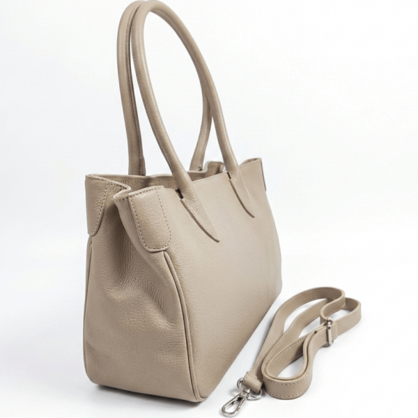 Елегантна бежова дамска чанта от естествена кожа, снимана под ъгъл на бял фон, с две изправени къси дръжки и допълнителна дълга дръжка със сребристи метални карабинери, поставена до нея.
