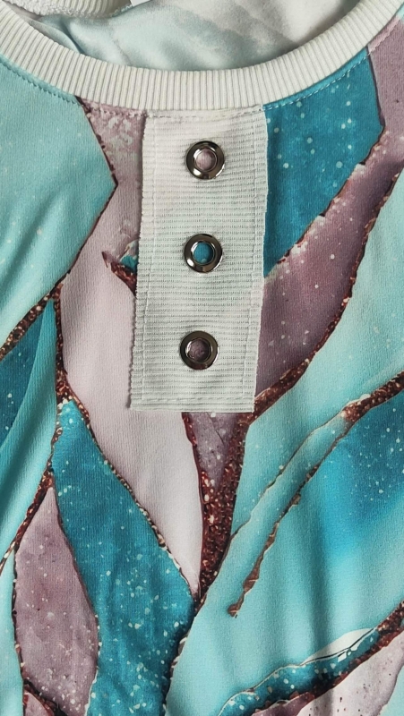 Едър план на деколтето и планката с три капси на туниката с абстрактен принт в тюркоаз, лавандулова и блестящи медни цветове.